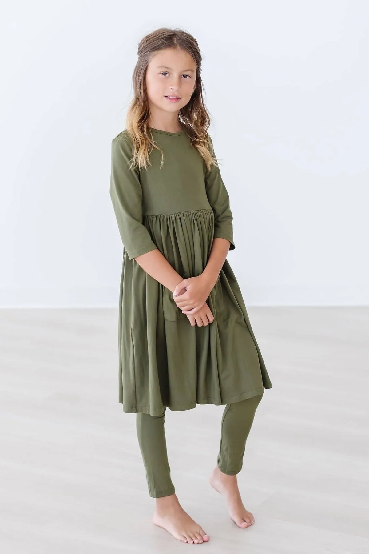 Girl wearing olive twirl dress for girls with three quarter sleeves standing barefoot indoors