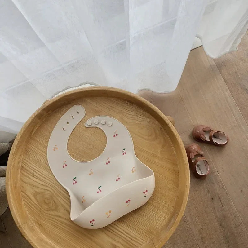 cream waterproof baby bibs with cherry pattern for infants displayed on wooden tray next to pink baby shoes