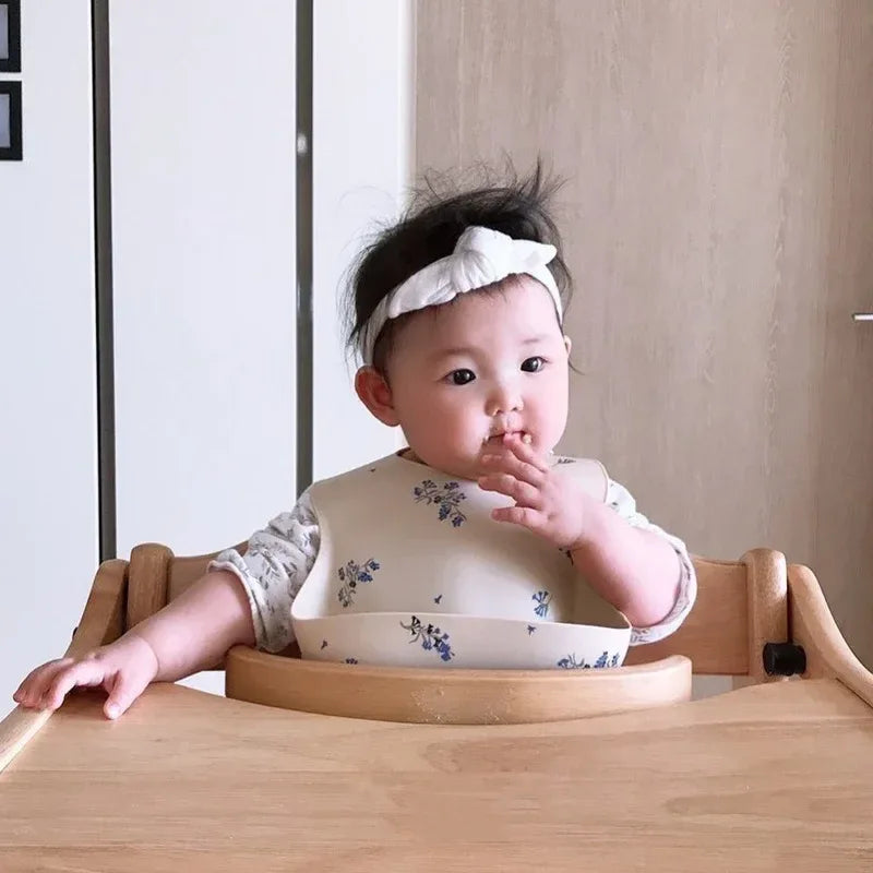 baby wearing cream waterproof baby bibs with blue floral print sitting in wooden high chair