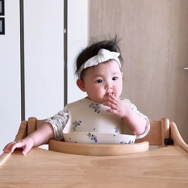 baby wearing cream waterproof baby bibs with blue floral print sitting in wooden high chair