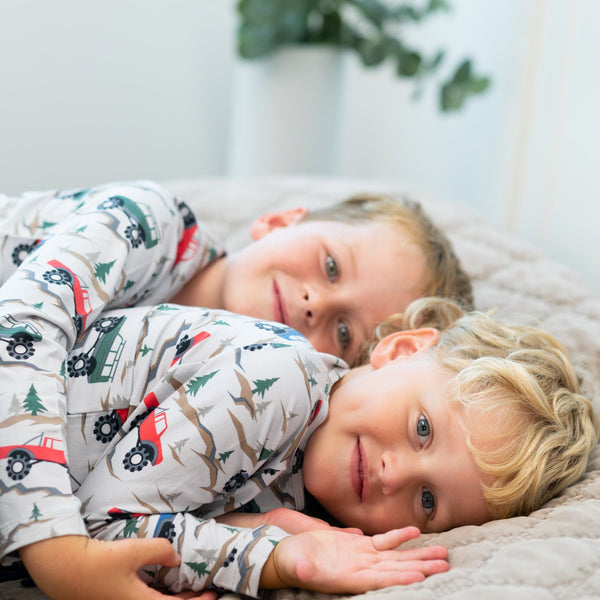 Two smiling boys lying on a bed wearing long sleeve henleys with a trucks and trees print, showcasing soft, comfortable button-up style for active play.