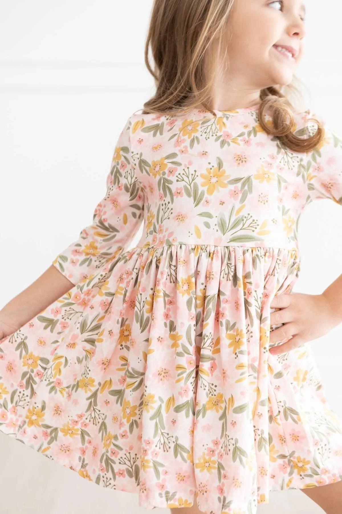 Young girl wearing peach floral girls twirl dress with 3/4 sleeves smiling and holding skirt