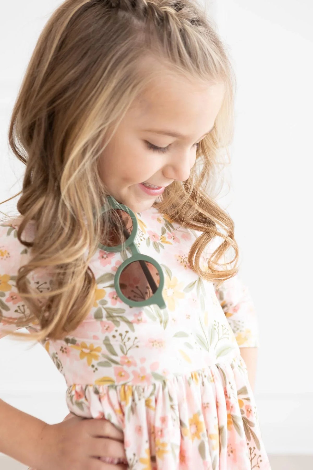 Young girl wearing peach floral girls twirl dress smiling and looking down with sunglasses hanging on dress