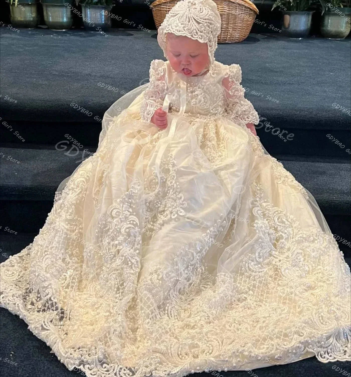 baby girl wearing an ivory pearl lace christening gown and matching bonnet sitting on dark carpeted stairs