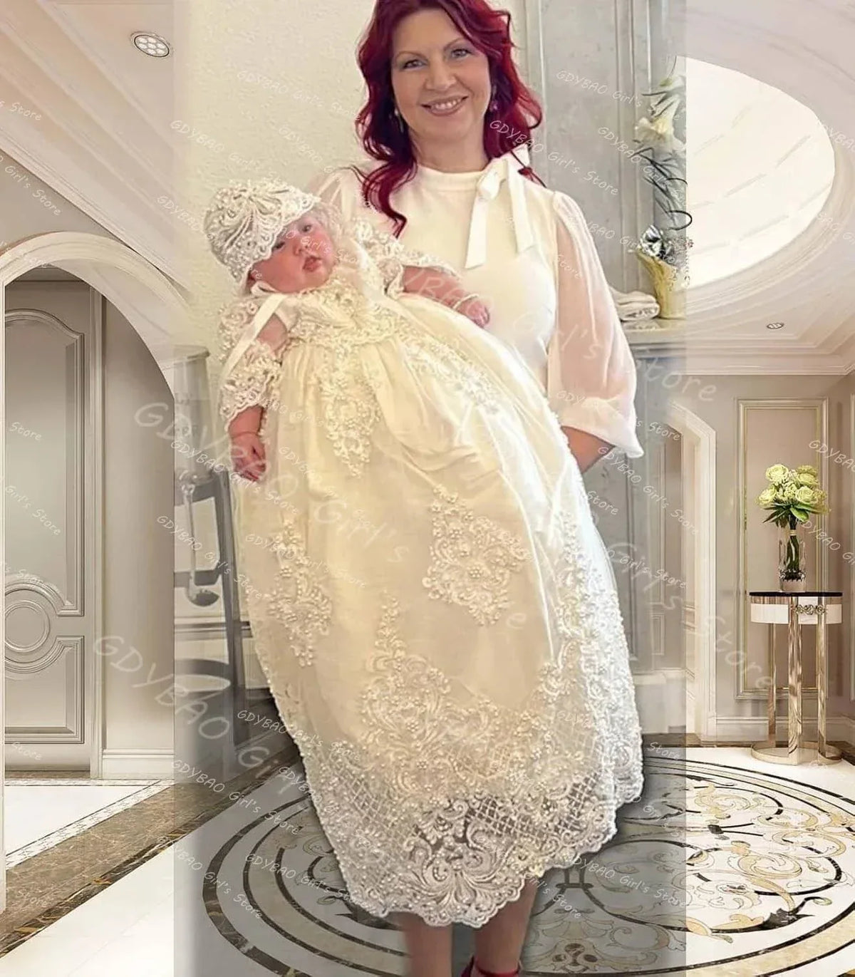 woman holding baby wearing white pearl lace christening gown and matching bonnet in elegant room