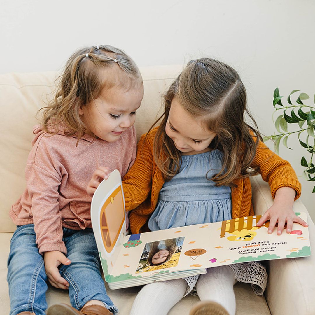 Two toddlers reading a colorful Peek A Photo interactive book sitting on a beige couch