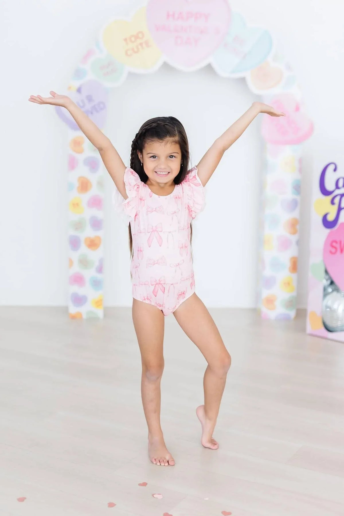 Girl wearing pink flutter sleeve baby leotard standing with arms raised smiling