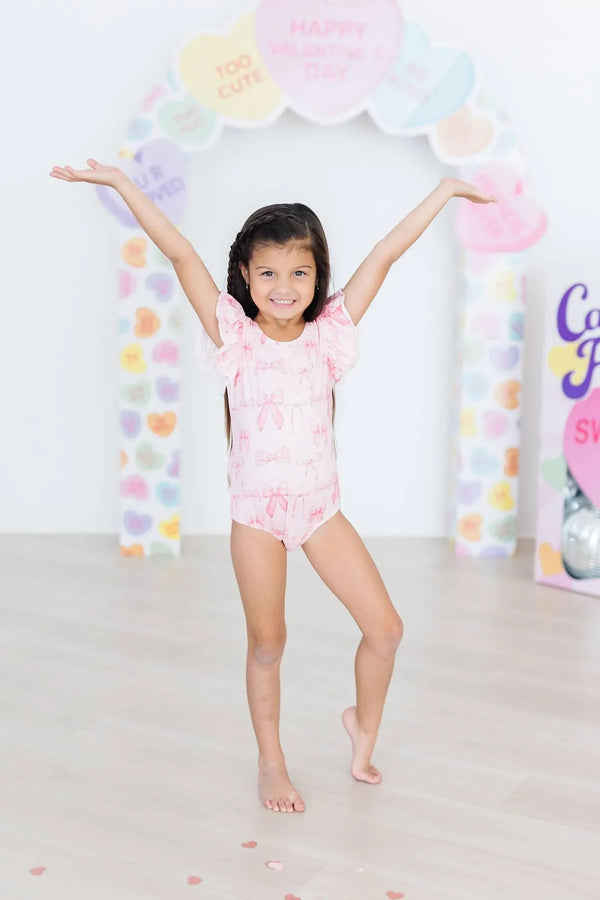 Girl wearing pink flutter sleeve baby leotard standing with arms raised smiling