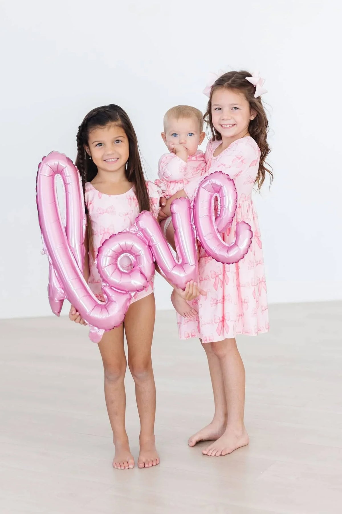 Toddler girl wearing pink flutter sleeve baby leotard holding pink love balloon with two girls