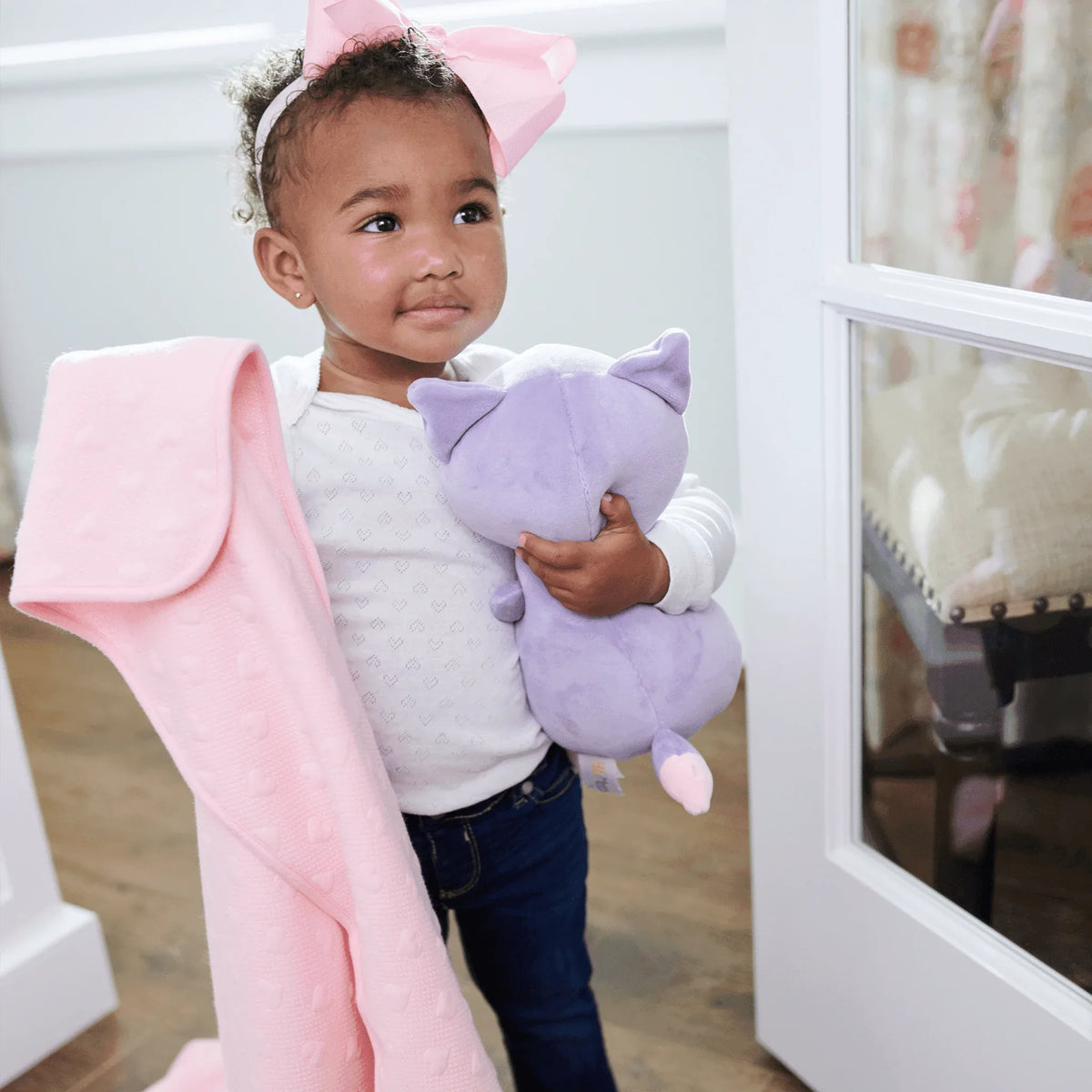 toddler girl holding pink knit baby blanket and purple stuffed cat toy standing indoors