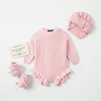 Pink knit gift set including ruffle romper, bow turban, and tie booties arranged on white surface