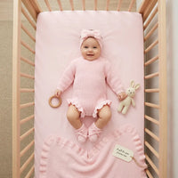Baby in pink knit romper wearing matching bow headband and booties in wooden crib with blanket