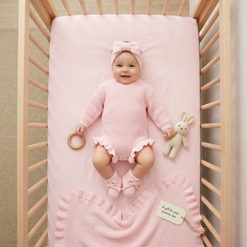 Baby in pink knit romper wearing matching bow headband and booties in wooden crib with blanket