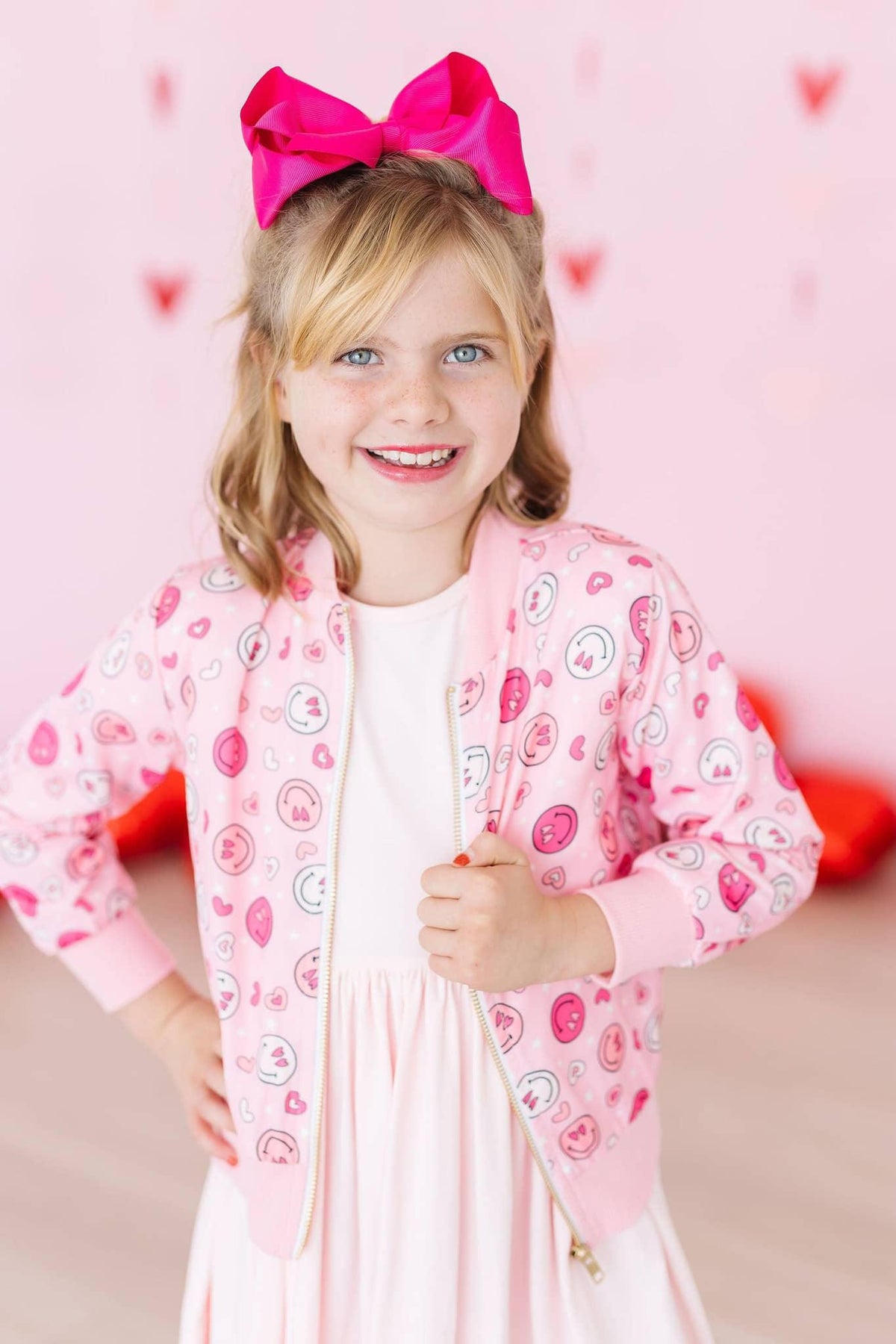 Girls pink satin jacket with heart pattern, long sleeve and zipper, worn by young girl with bow