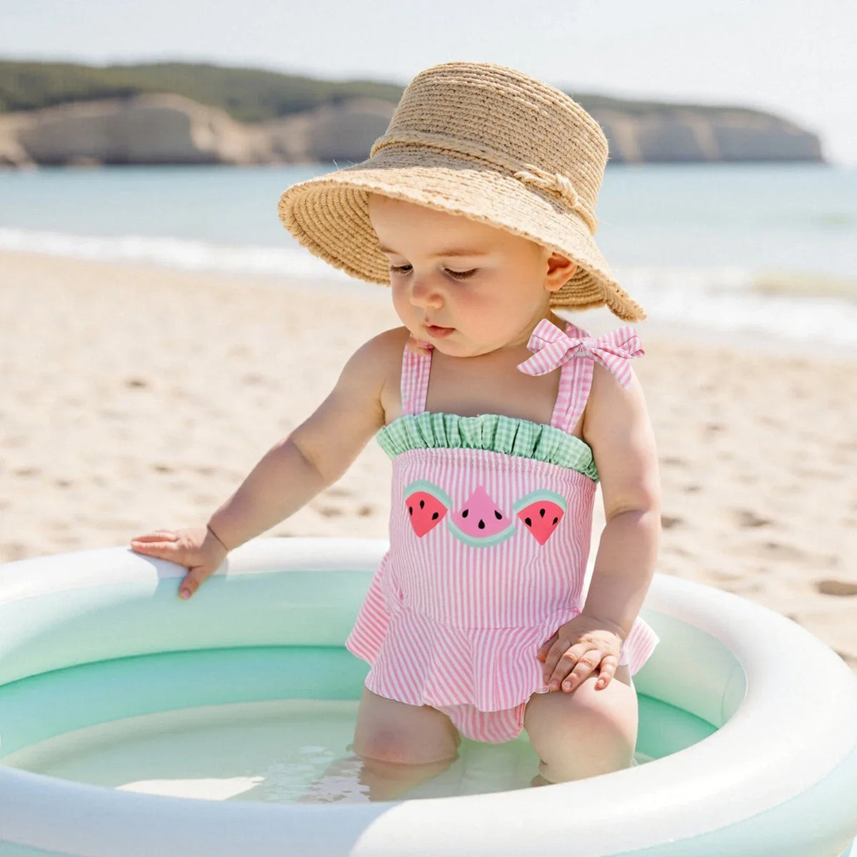 pink seersucker baby swimsuit worn by toddler girl with watermelon design and bow strap sitting in pool