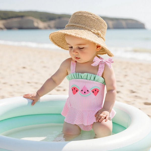 pink seersucker baby swimsuit worn by toddler girl with watermelon design and bow strap sitting in pool