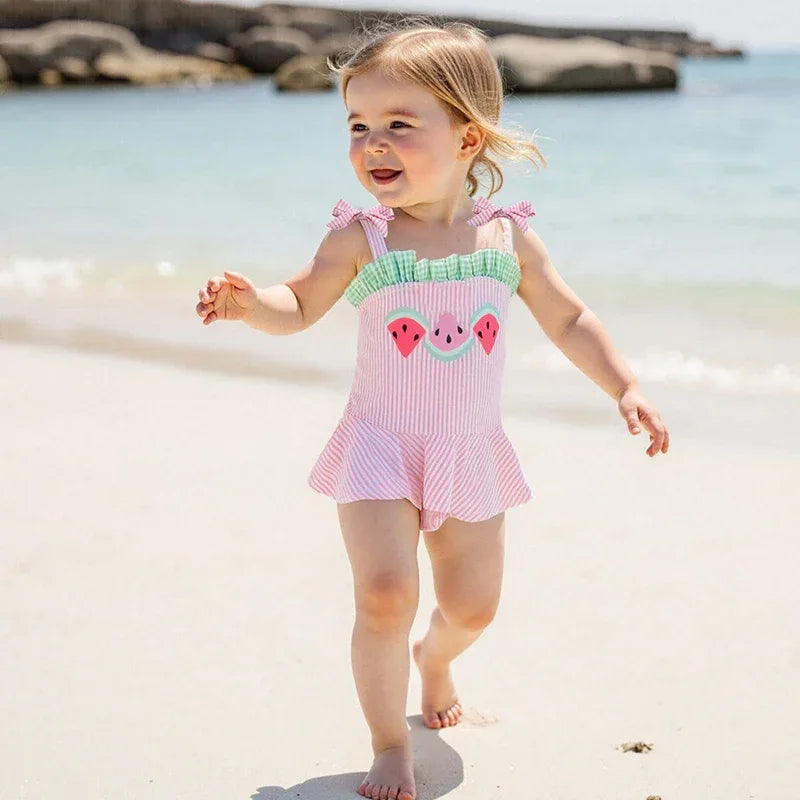 Toddler girl wearing a pink seersucker baby swimsuit with watermelon design and ruffle skirt at the beach