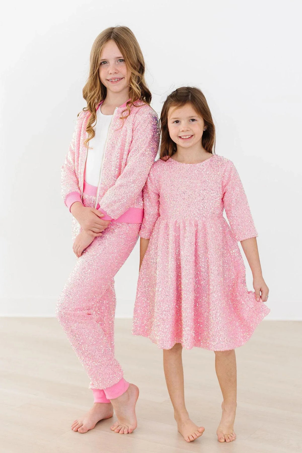 Two girls wearing pink sequin jacket for girls and matching pink sequin dress smiling and standing barefoot