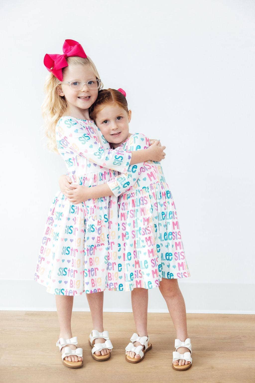 Two young girls wearing colorful pocket twirl dress hugging each other indoors