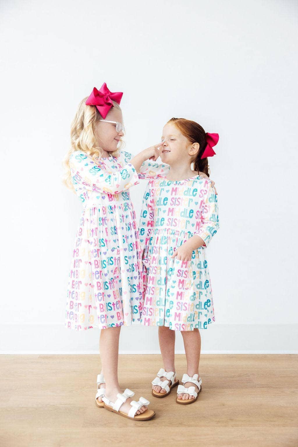 Two young girls wearing white pocket twirl dress with colorful letters and pink hair bows smiling and interacting