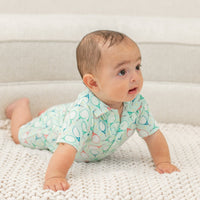 Baby wearing light green Polo Romper with tennis racket print, crawling on white knit blanket. Short sleeves and front buttons.
