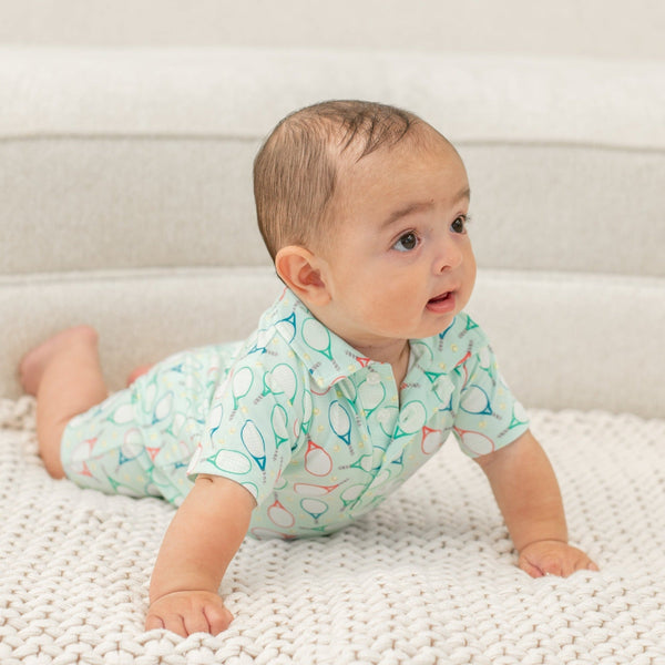 Baby wearing light green Polo Romper with tennis racket print, crawling on white knit blanket. Short sleeves and front buttons.