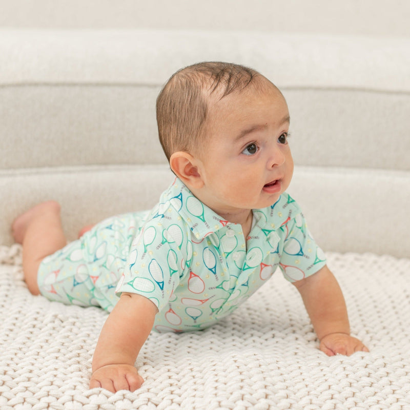 Baby wearing light green Polo Romper with tennis racket print, crawling on white knit blanket. Short sleeves and front buttons.