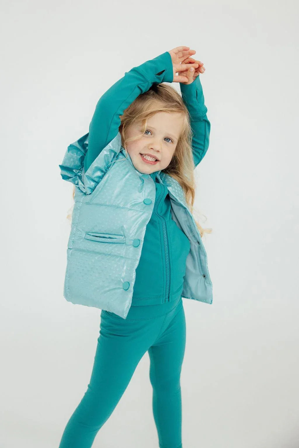 toddler girl wearing light blue puffer vest with ruffle detail over teal outfit