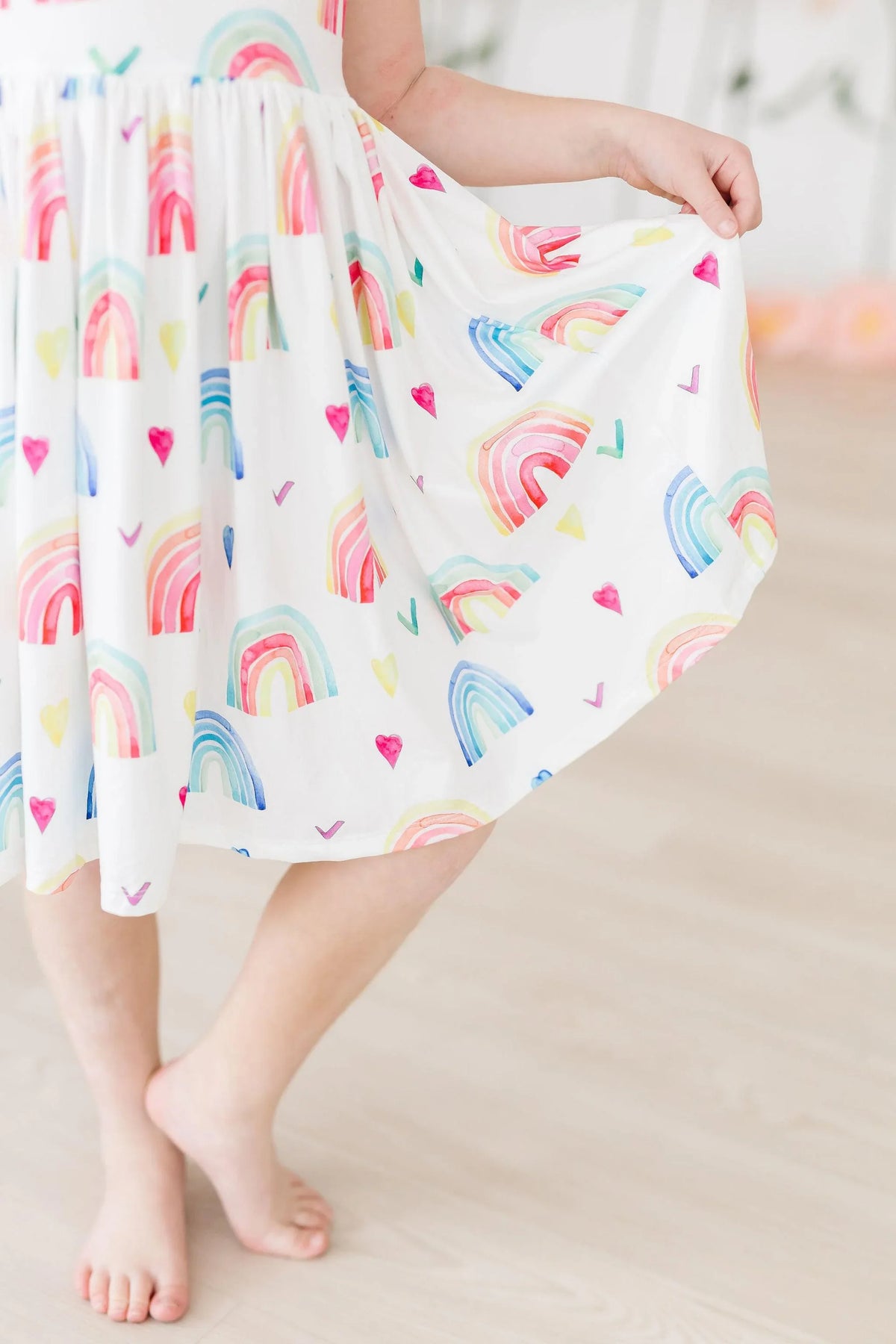 Young girl wearing white rainbow tank twirl dress for girls holding skirt while standing barefoot indoors