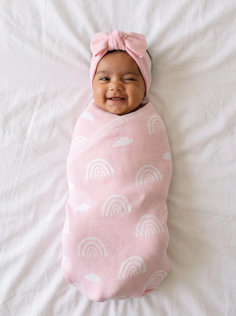 Smiling newborn in pink headband wrapped in pastel pink blanket with white rainbow and cloud jacquard pattern
