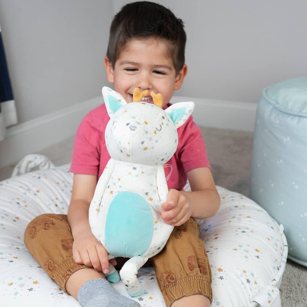 young boy holding white and blue reindeer plush toy for infants sitting on star patterned pillow