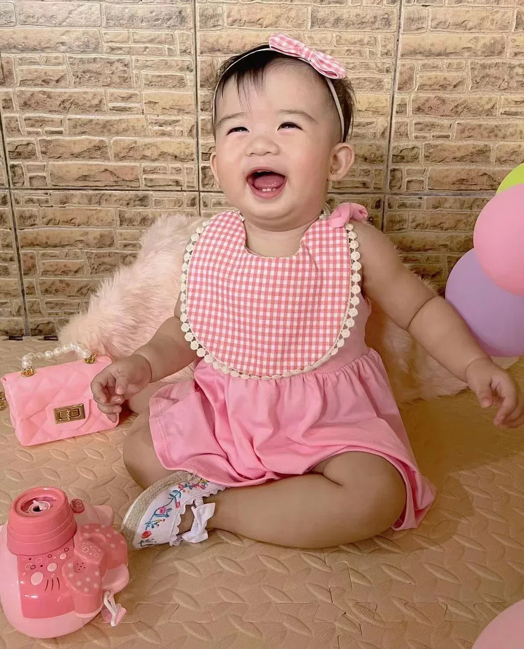 Smiling baby girl wearing pink dress and reversible baby bib and headband set sitting with toys