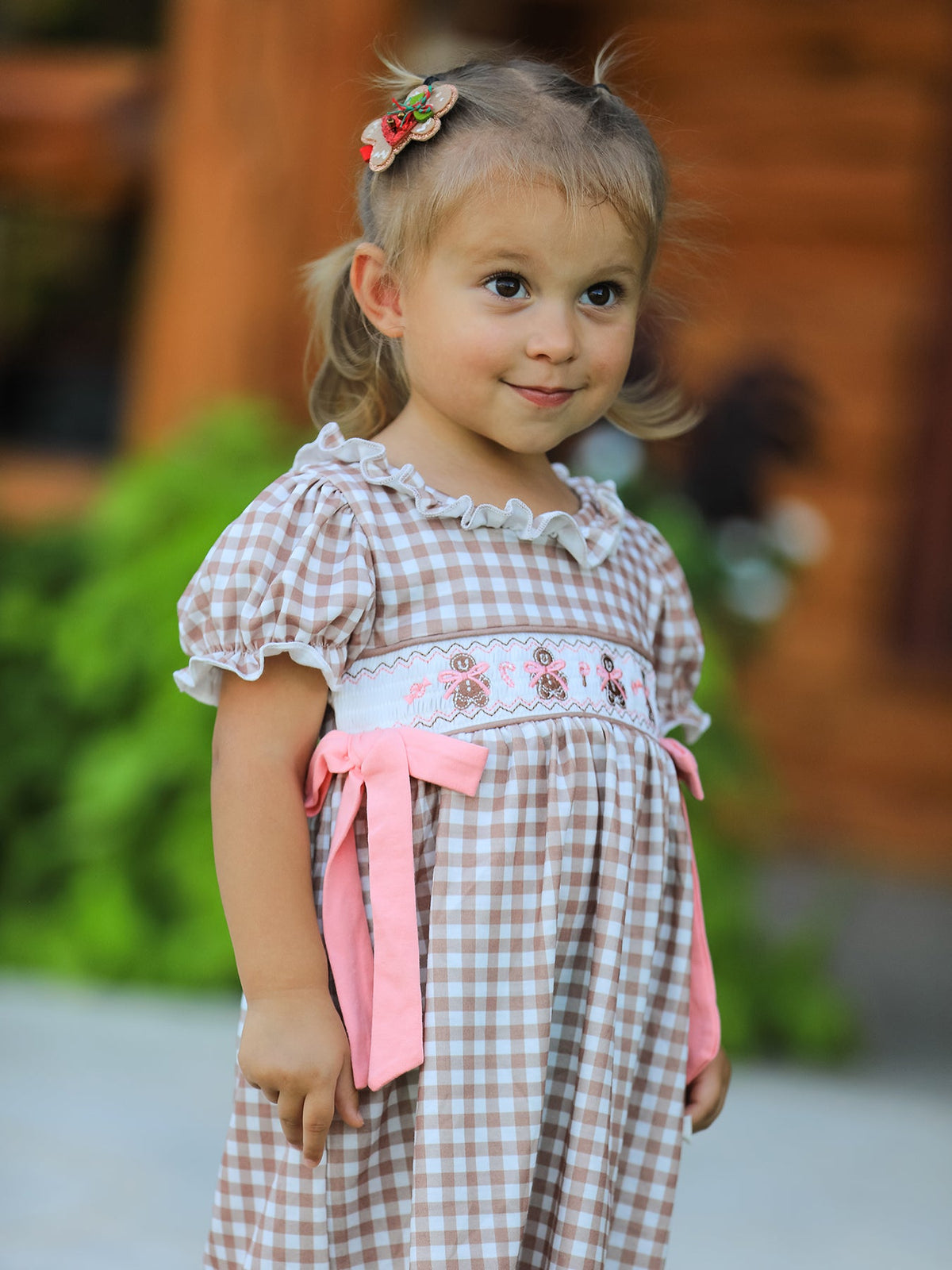 Little girl wearing Christmas vintage-style plaid dress with embroidered gingerbread and pink bows.