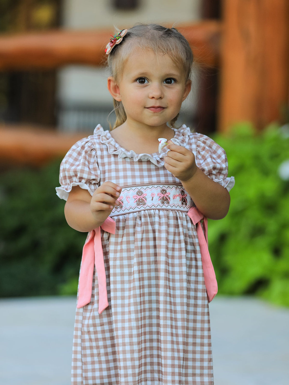 Little girl in Christmas vintage-style plaid dress with embroidered gingerbread and pink bows outdoors.