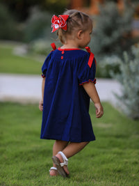 Toddler girl wearing navy Christmas dress with red bow shoulders and Santa sleigh hair bow, walking on grass.