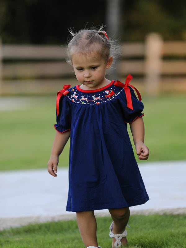 Toddler in navy Christmas dress with Santa sleigh embroidery and red ribbon tie shoulders.