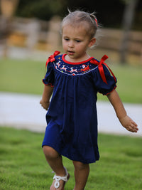 Toddler wearing navy Christmas dress with embroidered Santa sleigh, reindeer, and red ribbon shoulder ties. Festive holiday outfit for parties.