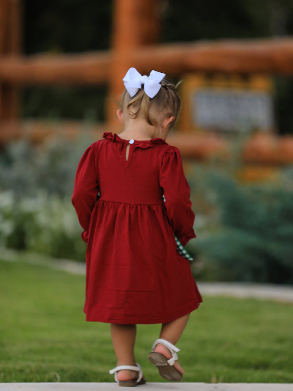 Back view of a girl in a red smocking embroidered dress with a green plaid bow and white hair bow.