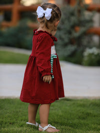 Back view of a girl wearing a red Christmas smocking embroidered dress with a green plaid bow and white hair bow.