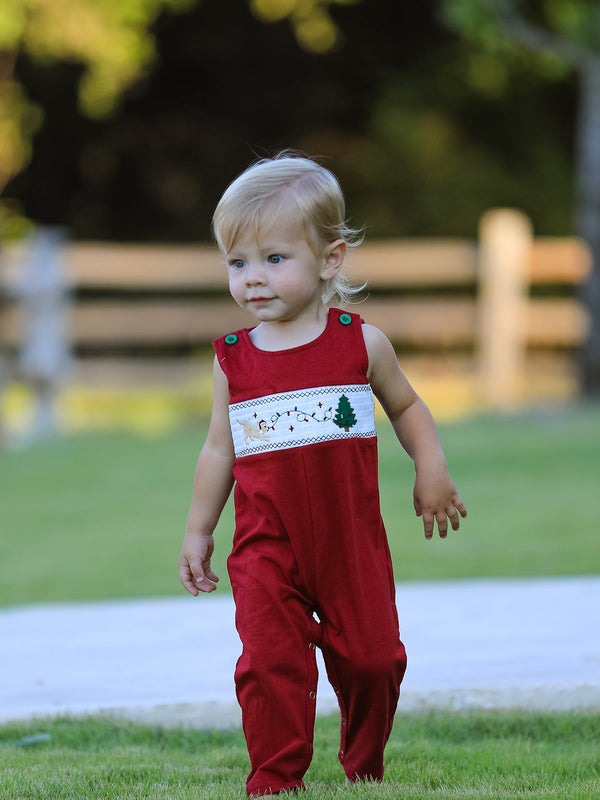 Toddler in red Christmas romper featuring smocked chest band with embroidered dog, tree, and lights.