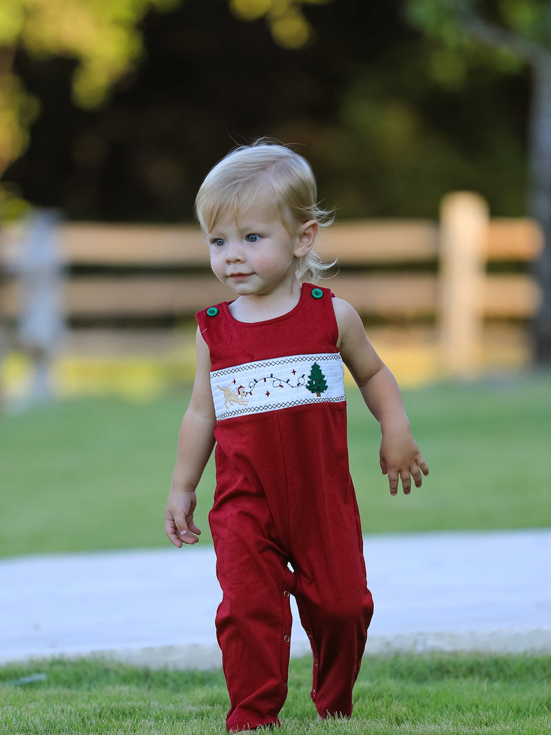 Toddler in red Christmas romper featuring smocked chest band with embroidered dog, tree, and lights.