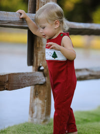 Baby boy in red Christmas Tree Dog smocking embroidered romper standing by a wooden fence. Festive holiday outfit with delicate stitching.