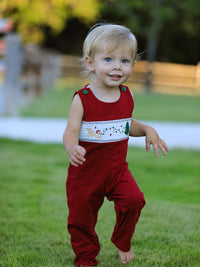 Baby boy in red romper with white smocking band featuring embroidered dog, Christmas tree, and lights. Festive holiday outfit.