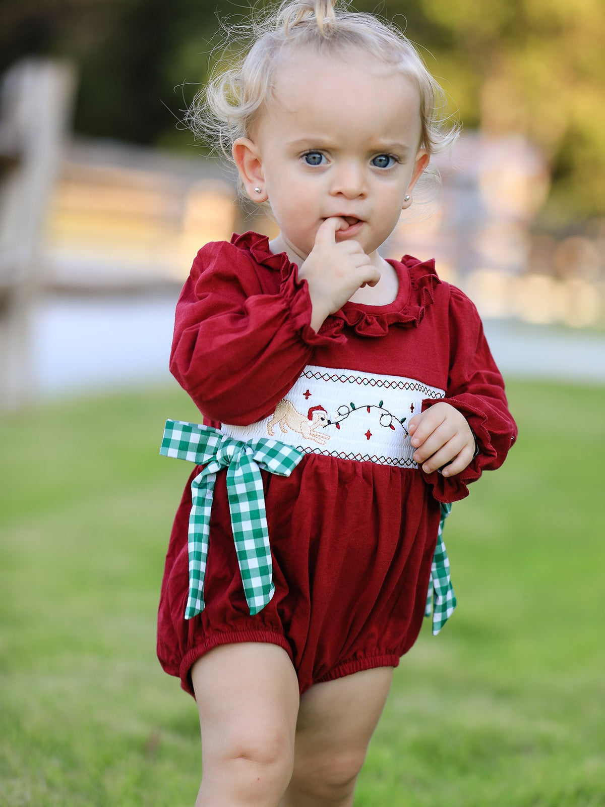 Baby girl wearing red Christmas romper with festive embroidered smocking and green plaid bow.