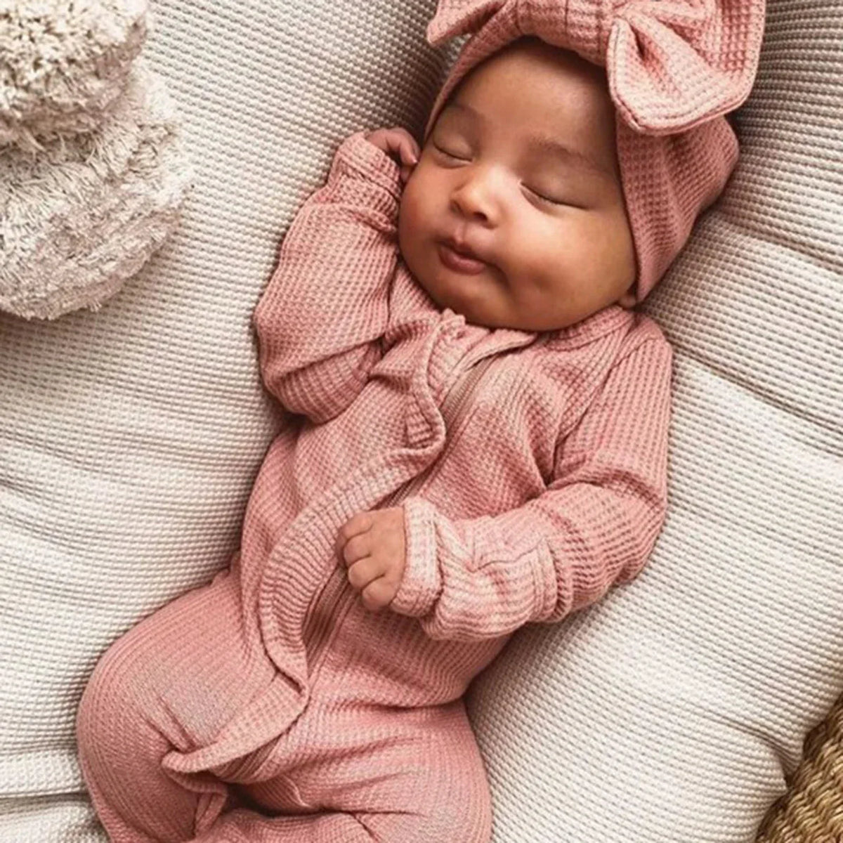 Sleeping newborn baby wearing a pink ribbed knit baby romper with headbow lying on a white cushion