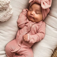 Sleeping newborn baby wearing a pink ribbed knit baby romper with headbow lying on a white cushion