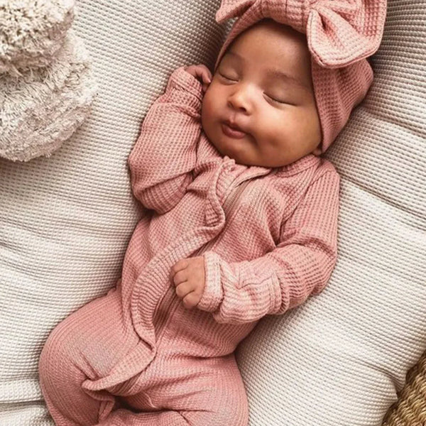Sleeping newborn baby wearing a pink ribbed knit baby romper with headbow lying on a white cushion