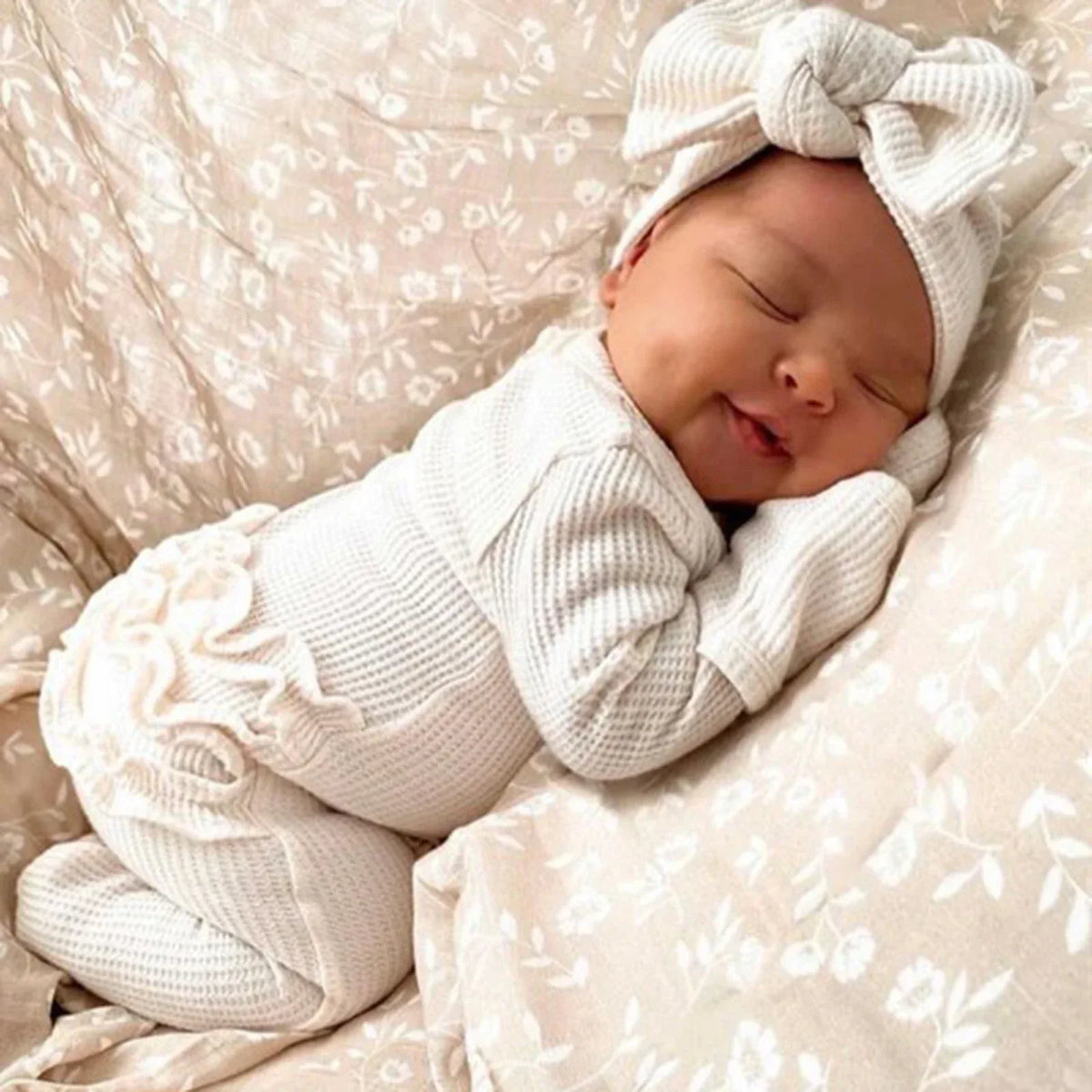newborn baby girl sleeping in cream ribbed knit baby romper with headbow on floral bedding
