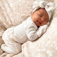 newborn baby girl sleeping in cream ribbed knit baby romper with headbow on floral bedding