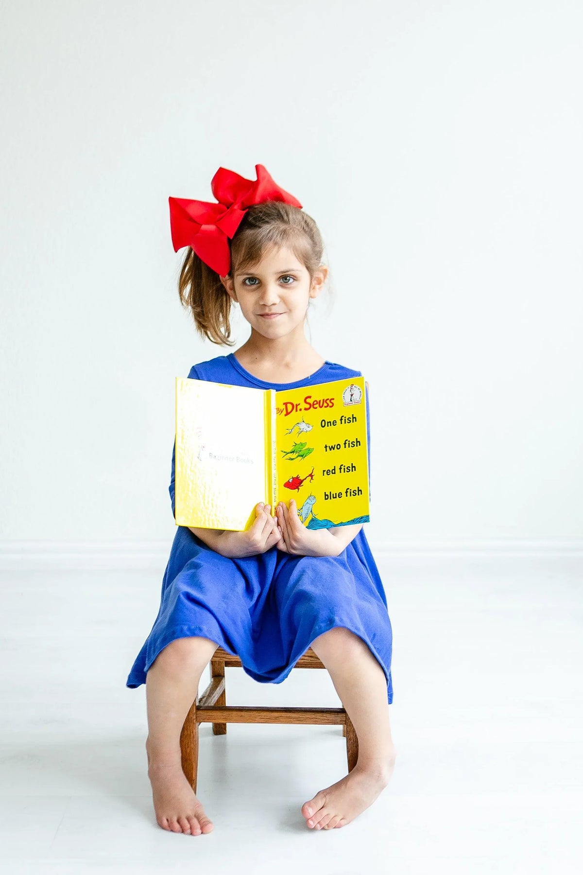 Royal blue twirl dress for girls, 3 4 sleeve pocket dress worn by young girl with red bow reading a Dr Seuss book